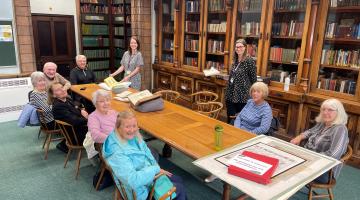 A group of older people sitting around a long wooden table surrounded by cases of books